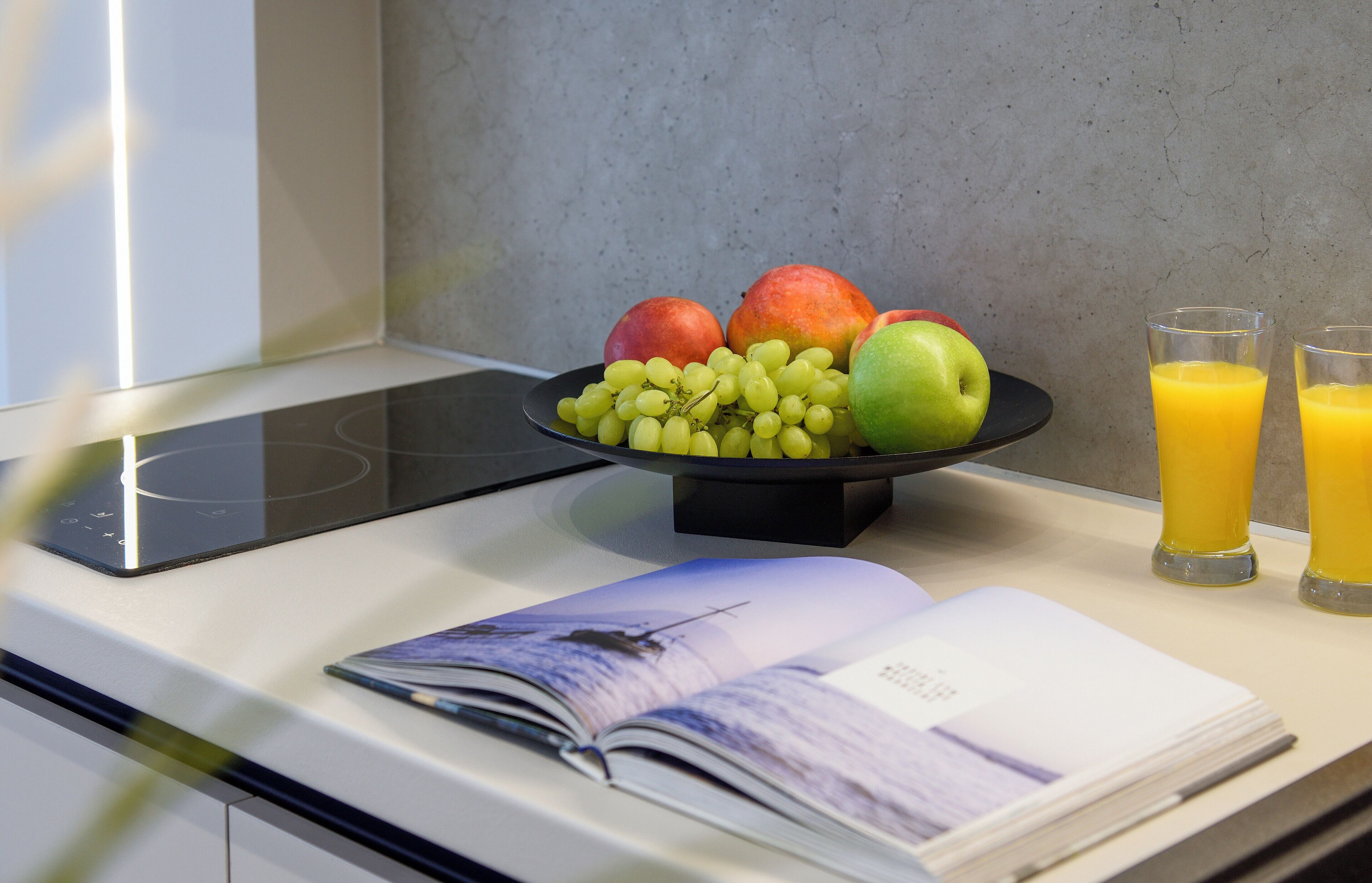 Una encimera de cocina con un frutero que contiene manzanas y uvas, dos vasos de jugo de naranja y una revista.