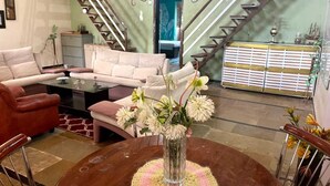 A living room with a sofa, armchairs, a coffee table, and a vase of flowers. There is a staircase with a railing and a cabinet with decorative items.