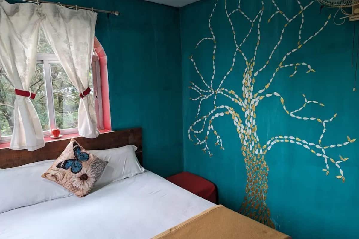 A bedroom with a bed, a window with curtains, a red bench, and a wall mural of a tree.