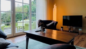 A living room with a sofa, a coffee table, a TV, and a sliding glass door leading to an outdoor area with chairs and greenery. - Traumhafte Ferienwohnung am Rande der Lüneburger Heide im Celler Land (Adelheidsdorf)