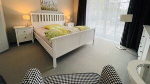 A neatly arranged bedroom with a bed, bedside tables, a lamp, a chair, and a view of the outdoors through a window. - Traumhafte Ferienwohnung am Rande der Lüneburger Heide im Celler Land (Adelheidsdorf)