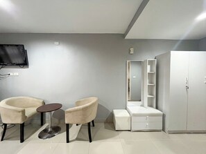 A modern room with a flat-screen TV, two armchairs, a small round table, a vanity with a mirror and shelves, and a large wardrobe.