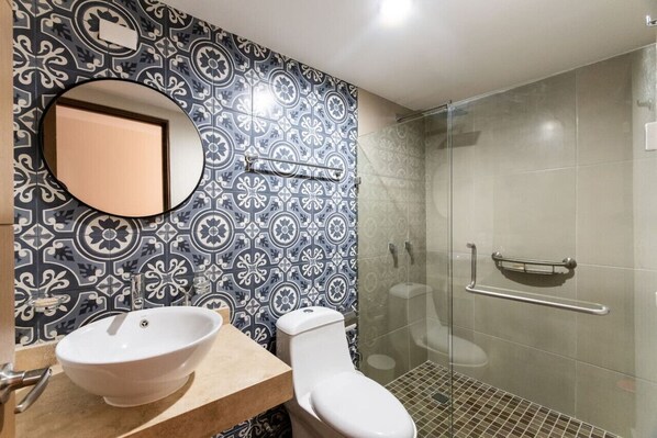 A bathroom with a patterned tile backsplash, a round mirror, a sink, a toilet, and a glass shower enclosure.