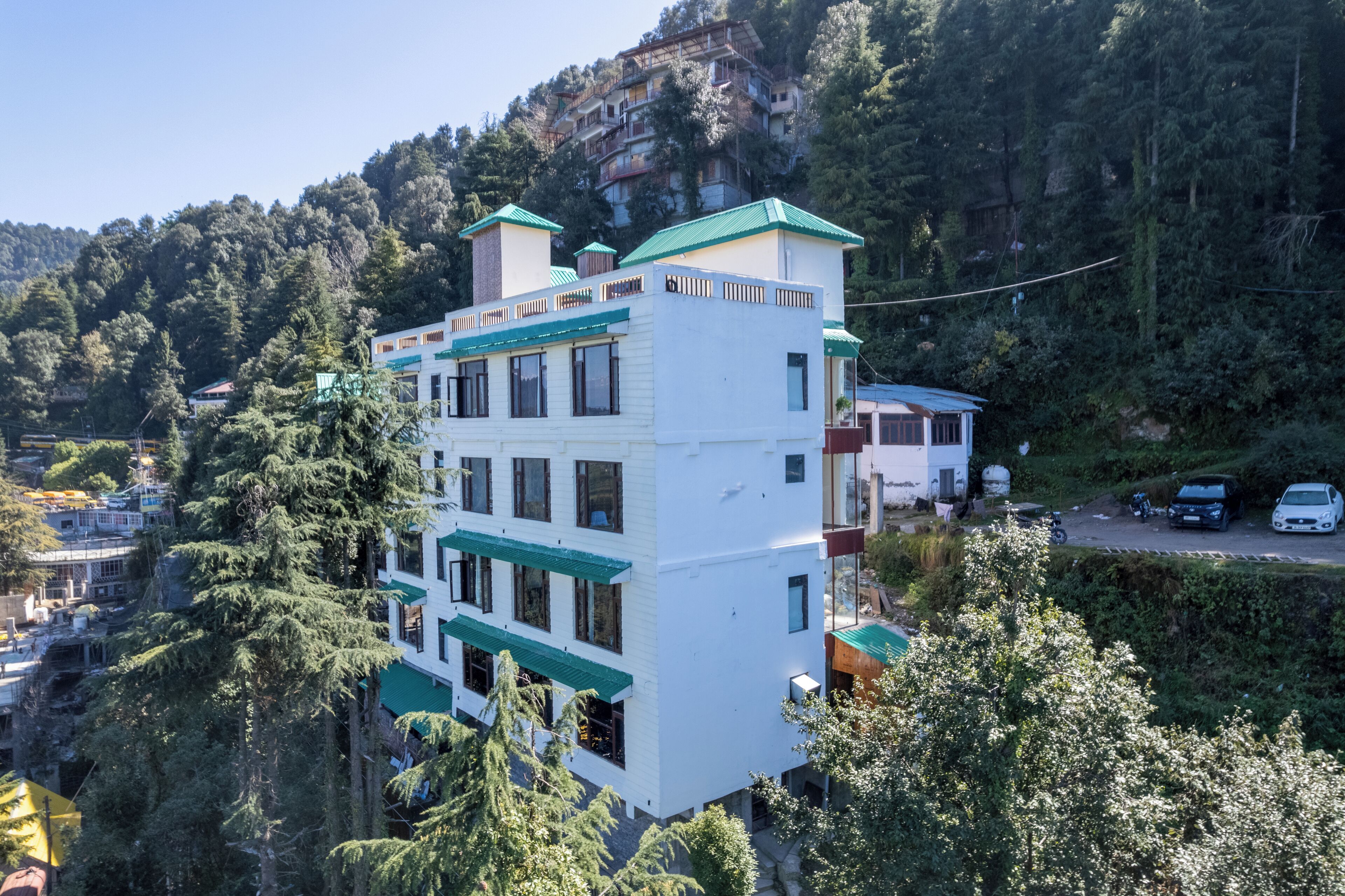 A multi-story building with a white facade and green roof, surrounded by dense forest and hills.