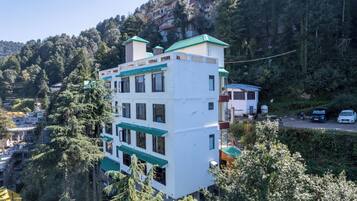 A multi-story building with a white facade and green roof, surrounded by dense forest and hills.