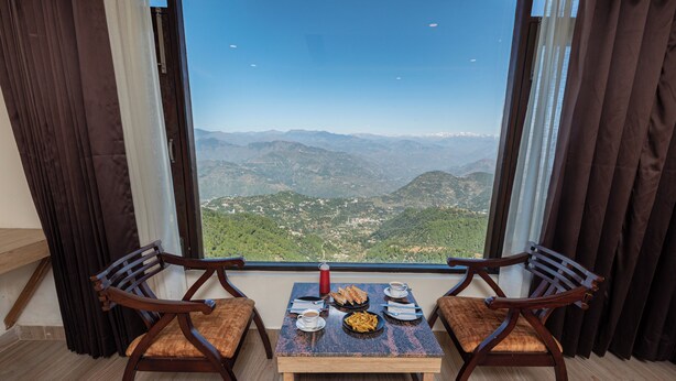 Un comedor con dos sillas de madera, una mesa pequeña y vistas a las montañas a través de un ventanal grande.