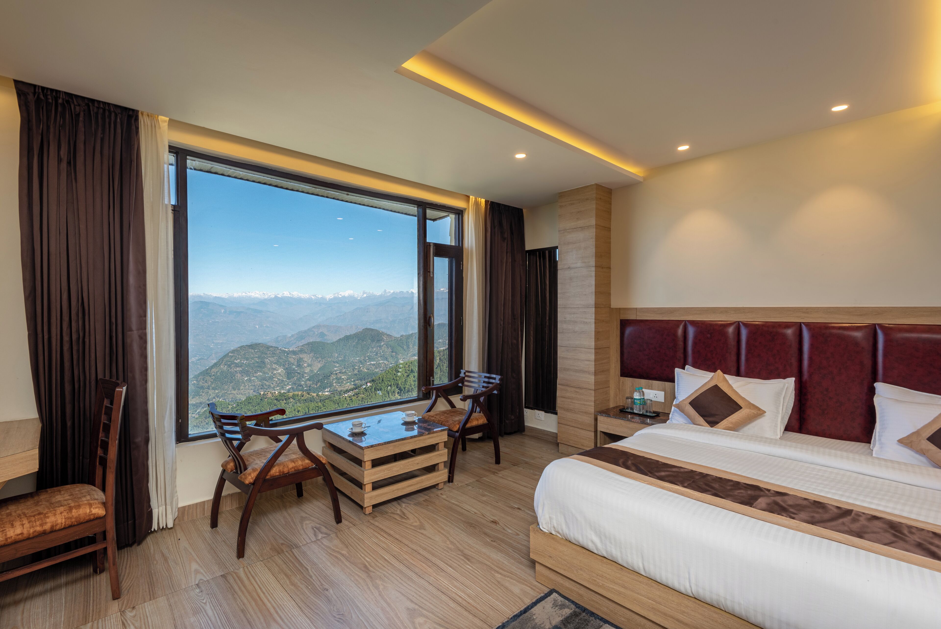 A hotel room with a large window offering a mountain view, a bed with a red headboard, a wooden desk, and a chair.
