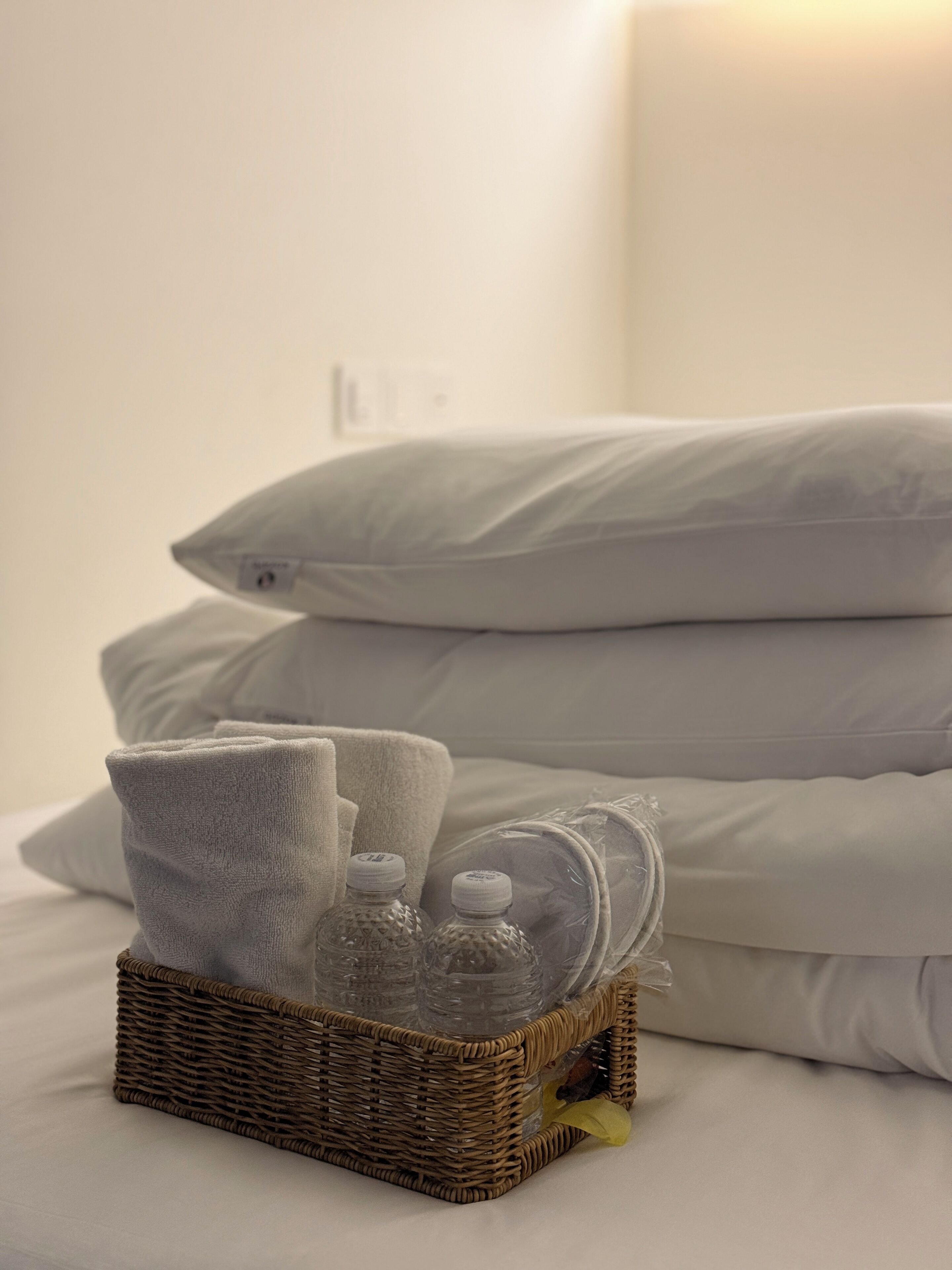 A neatly made bed with white bedding and pillows, a wicker basket with toiletries, and a wall socket.