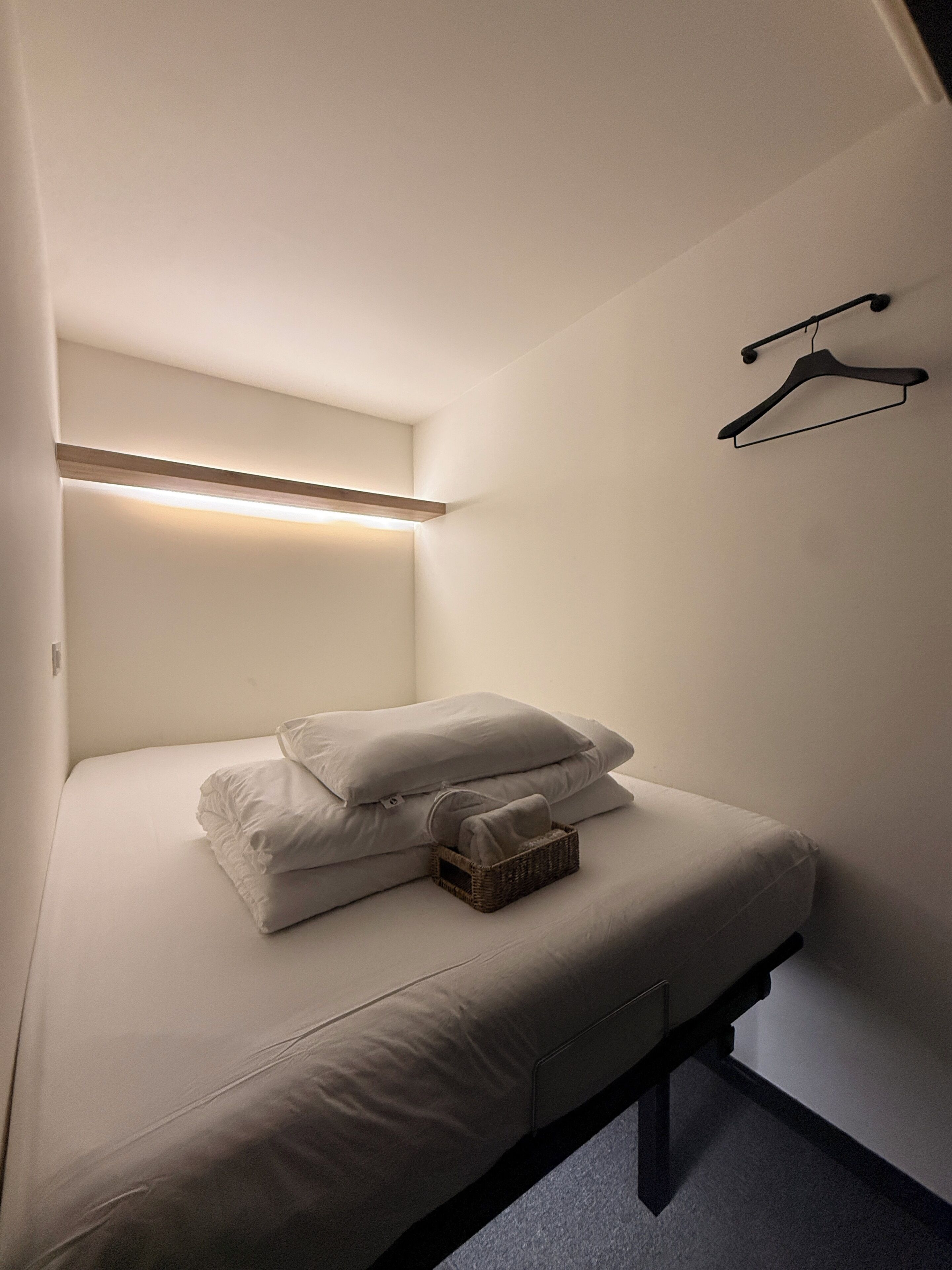 Quarto pequeno com cama de solteiro, mesa de cabeceira, luminária suspensa e suporte de parede para roupas.