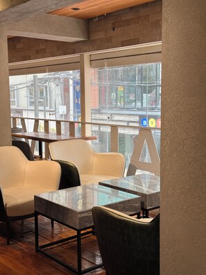 A seating area with a sofa and tables, large windows with a view of the street, and a brick wall. - KOZAYO Hongdae Hostel (Seoul)