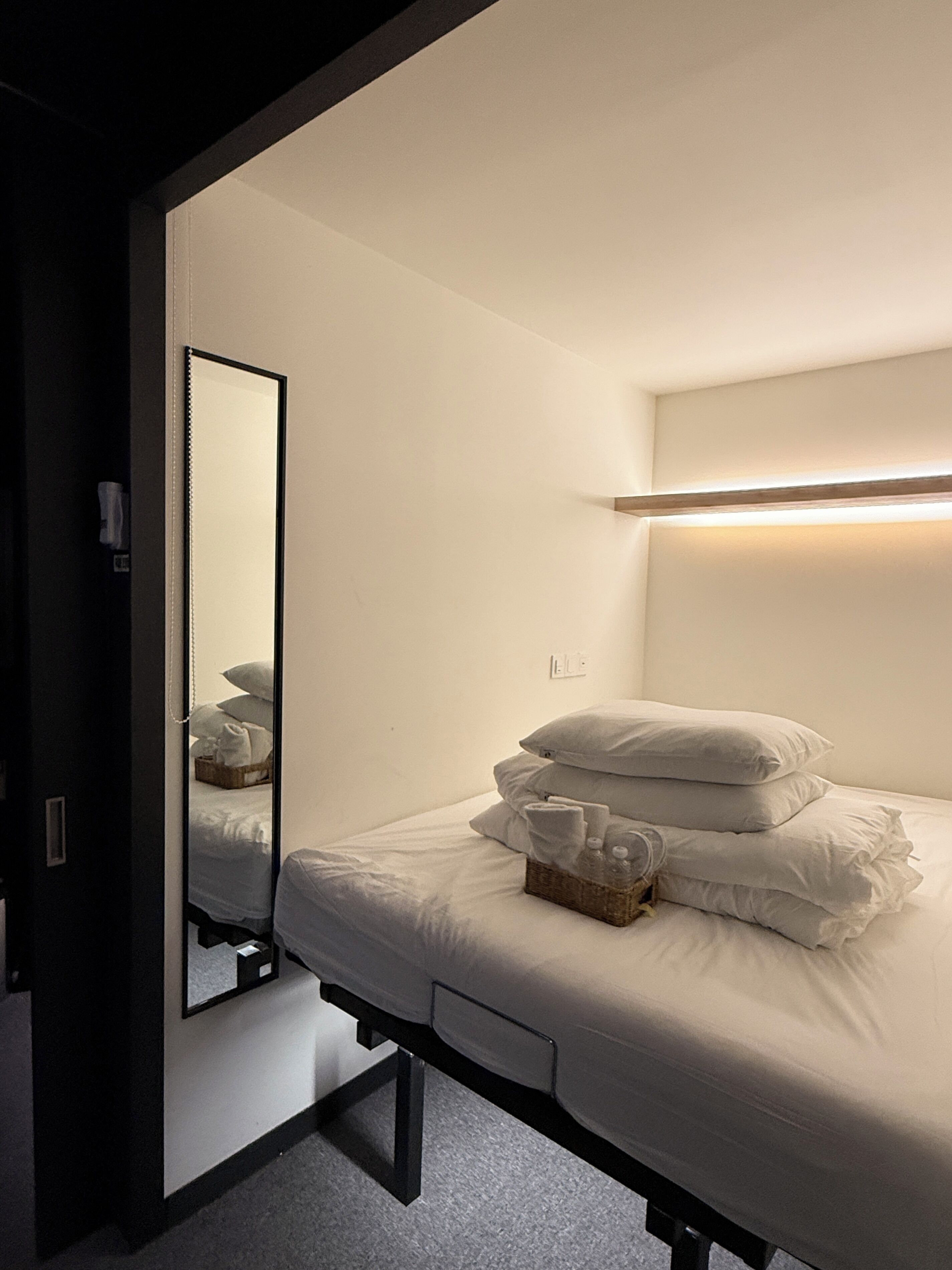 A single bed with white bedding and pillows, a bedside table with a basket, and a mirror reflecting the bed.