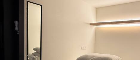 A single bed with white bedding and pillows, a bedside table with a basket, and a mirror reflecting the bed.