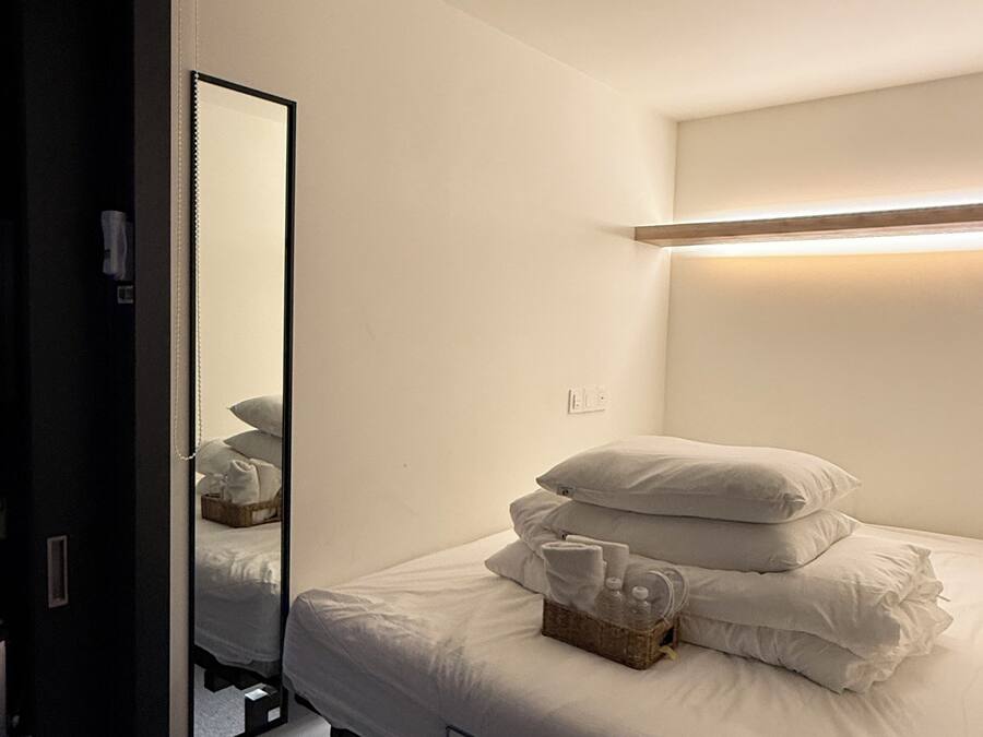 Una cama individual con ropa blanca y almohadas, una mesita de noche con una cesta, y un espejo que refleja la cama.