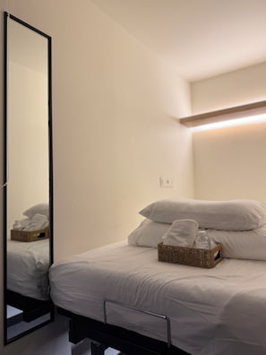Un lit bien fait, avec du linge de lit blanc, un panier en osier contenant une bouteille et un miroir qui reflète un coin de la pièce.