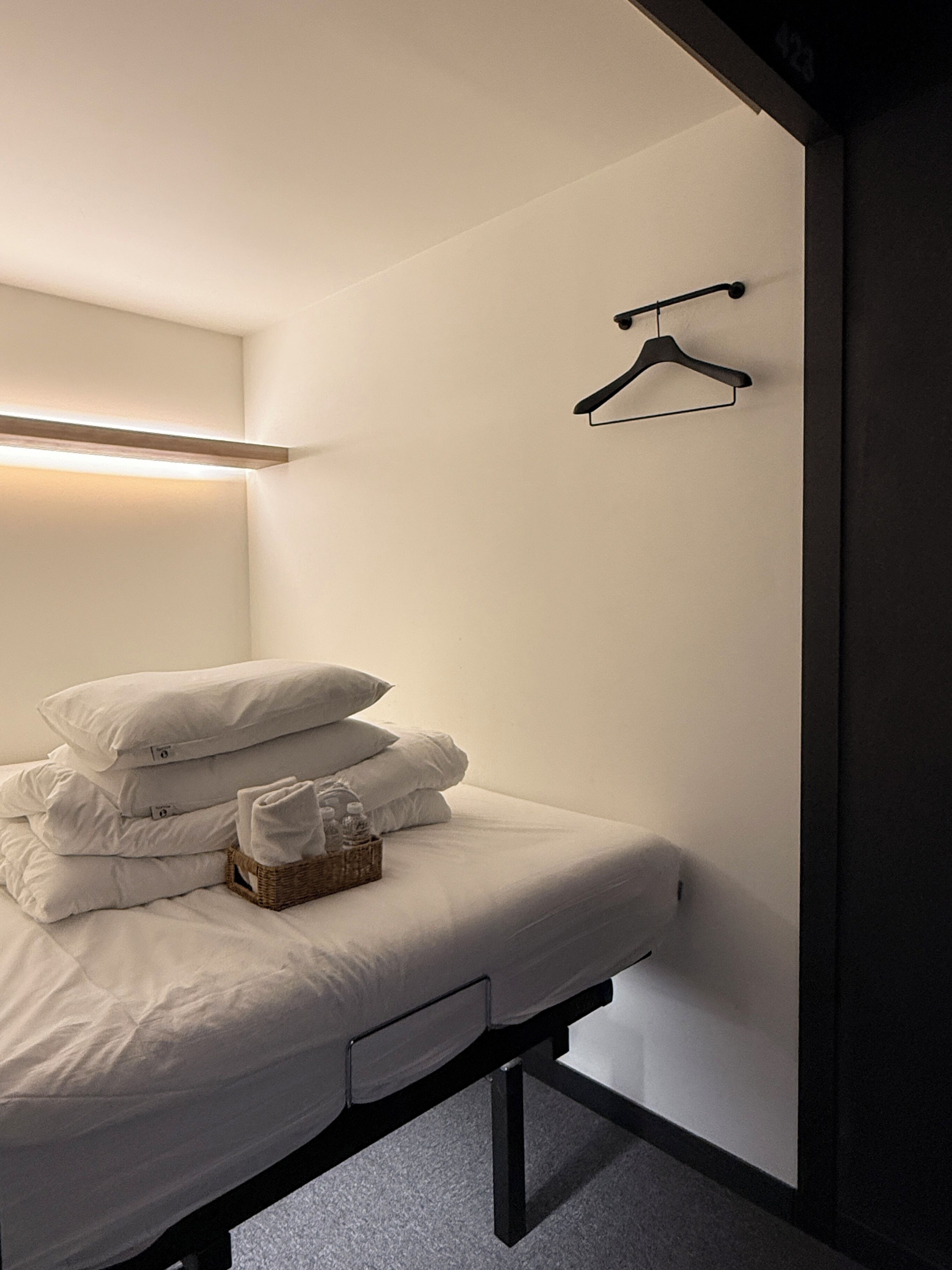 A neatly made bed with pillows and a basket of toiletries, a wall-mounted hanger, and a bedside table with drawers.