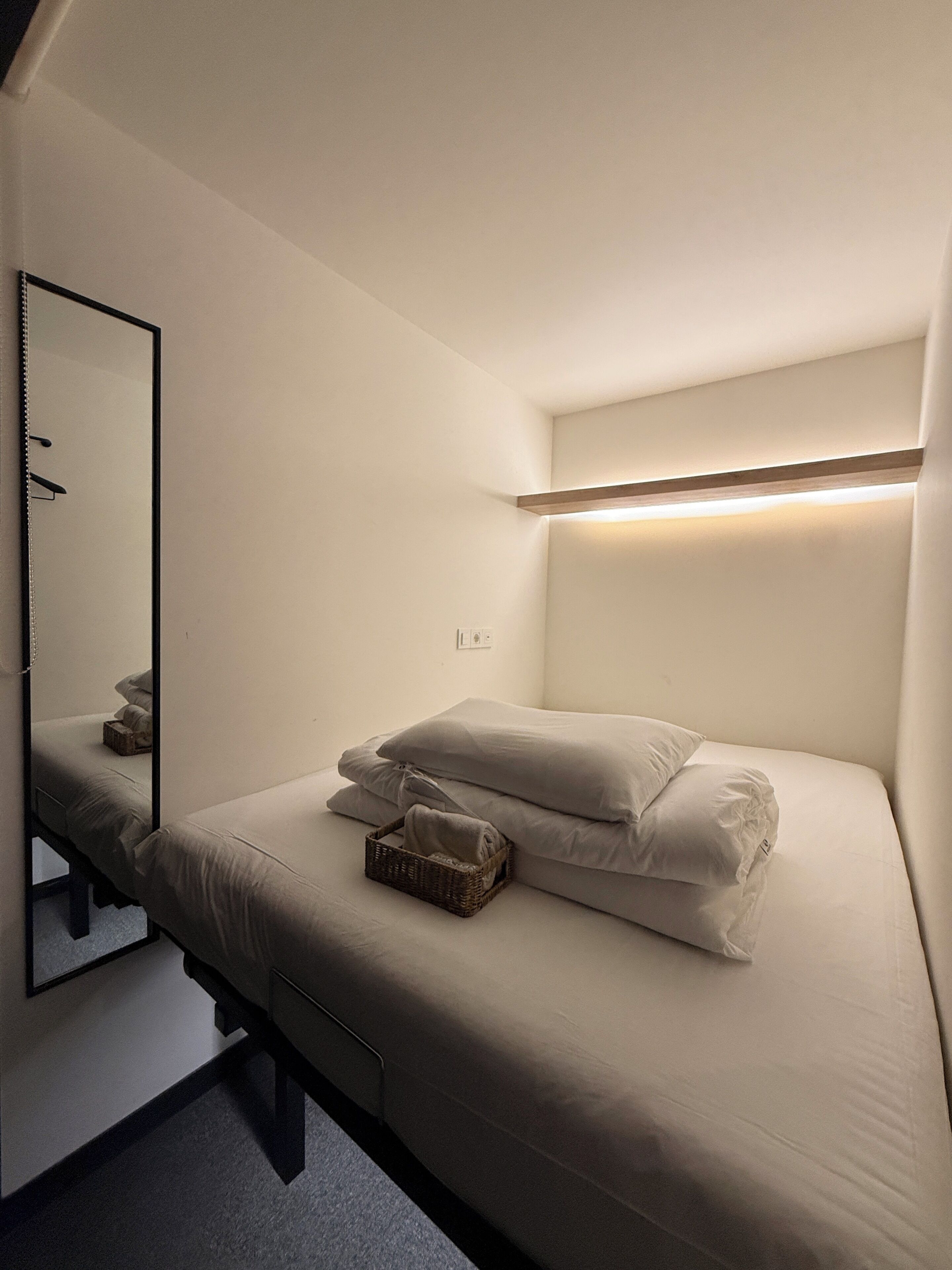 A single bed with pillows and a basket, a bedside table, a mirror, and a wall-mounted light fixture.