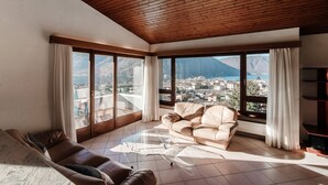 A spacious living room with a wooden ceiling, large windows offering a view of a mountainous landscape, a beige sofa, and a brown leather couch. - Flat in the Gulf of Melano (Miralago) (Val Mara)