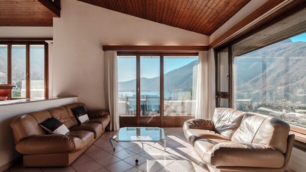 A modern living room with leather sofas, a glass coffee table, and large windows offering a mountain view. - Flat in the Gulf of Melano (Miralago) (Val Mara)