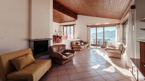 A spacious living room with a fireplace, leather sofas, and large windows offering a view of the mountains. - Flat in the Gulf of Melano (Miralago) (Val Mara)