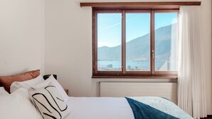 A bedroom with a large window, a bed with white bedding, and a view of mountains and a lake. - Flat in the Gulf of Melano (Miralago) (Val Mara)