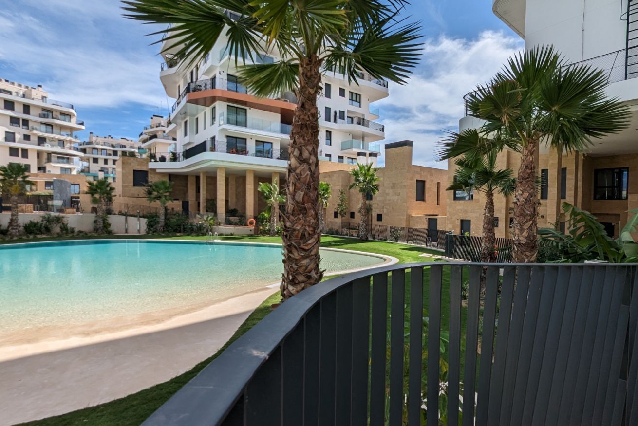A poolside view of a residential building with multiple balconies, palm trees, and a clear blue sky.