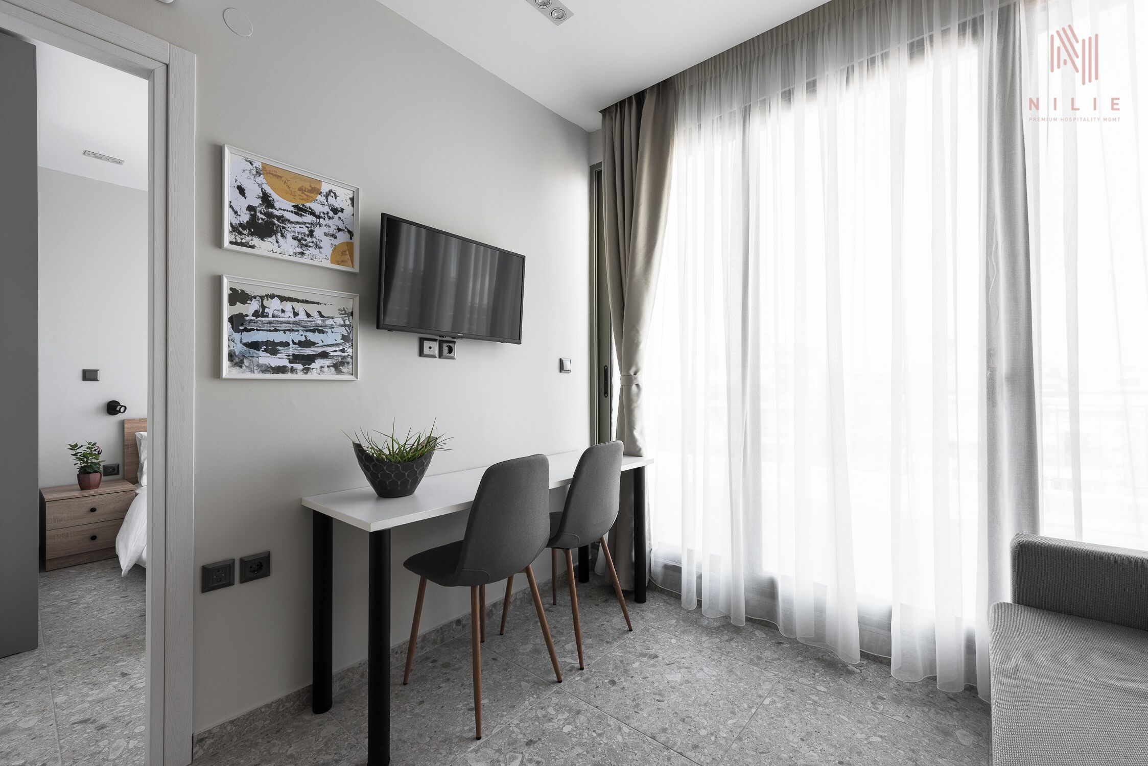 Habitación de hotel moderna con una televisión de pantalla plana montada en la pared, una mesita con dos sillas y un ventanal con cortinas de gasa.