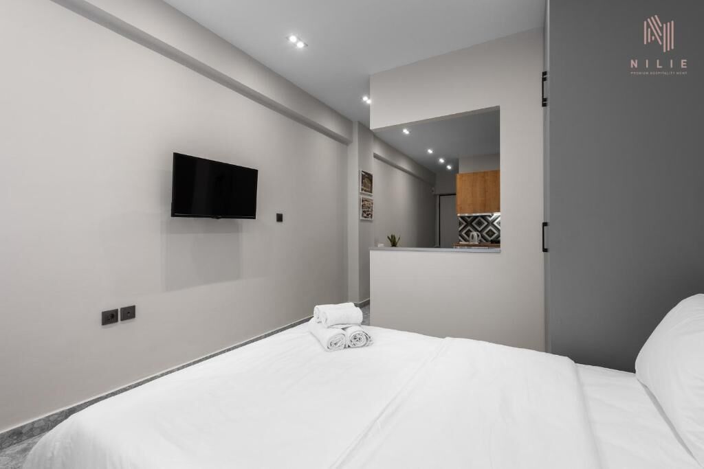 Un hotel moderno con una televisión de pantalla plana montada en la pared, una cama bien hecha con sábanas blancas y una pequeña planta en la mesita de noche.
