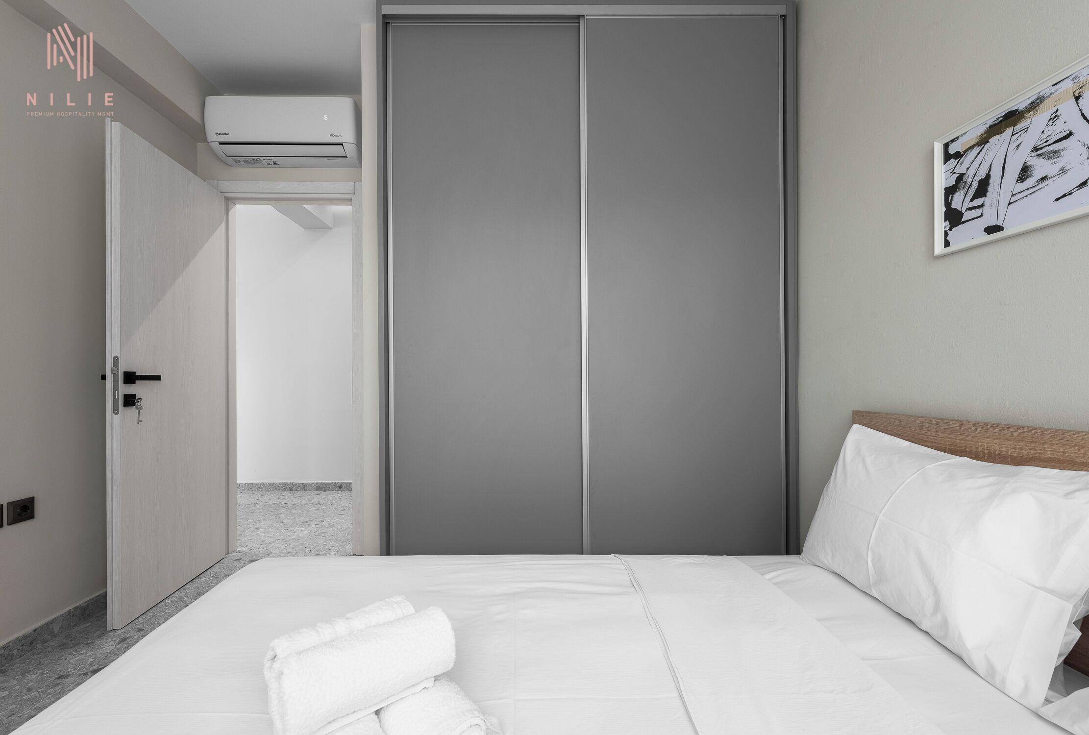 Una habitación de hotel con cama, ropa de cama blanca, un armario cerrado y un equipo de aire acondicionado en la pared.