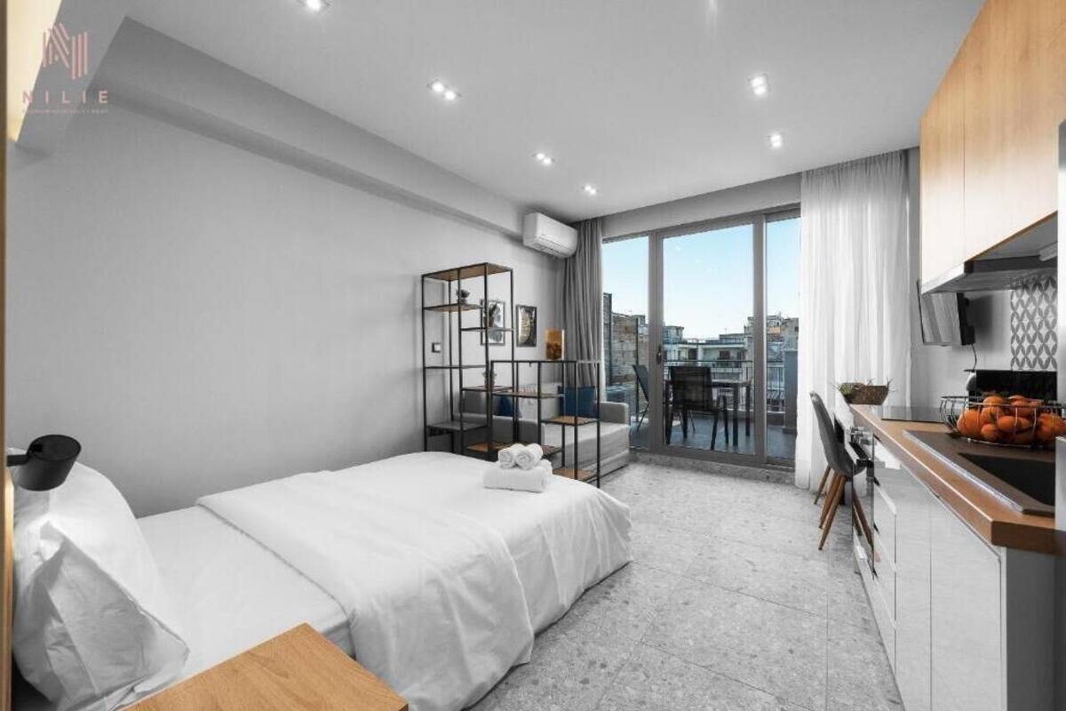 A modern hotel room with a bed, a desk with a sink, and a balcony with a city view.