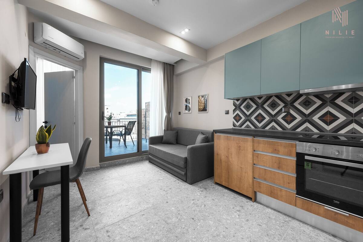 A modern kitchen with a built-in oven, a dining area with a table and chairs, a sofa, and a large sliding glass door leading to a balcony.