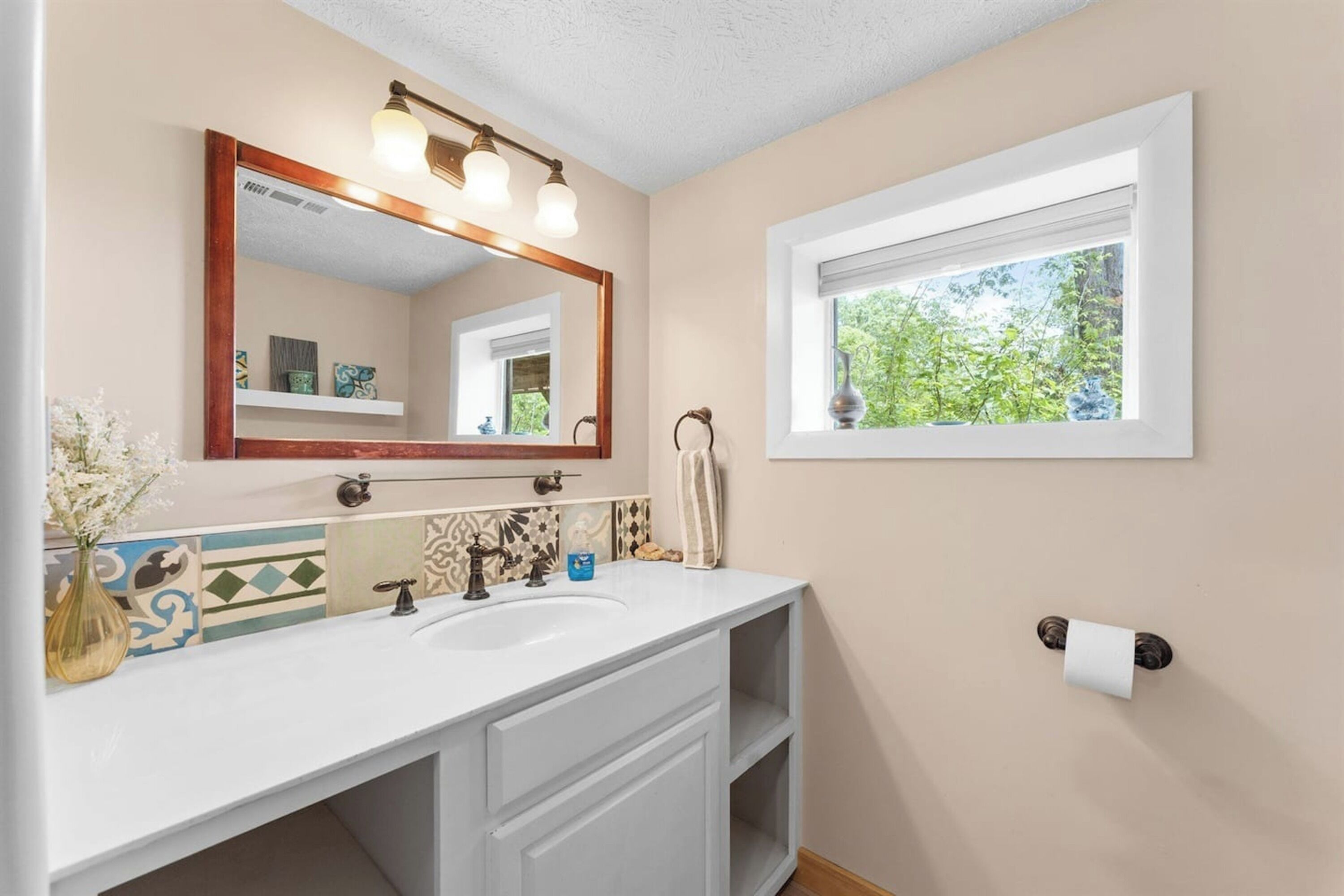 A bathroom with a white sink, a mirror with a wooden frame, a window with a view of greenery, and a towel rack.