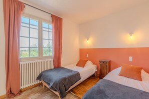 A bedroom with two beds, a window with curtains, a radiator, and a wooden nightstand.