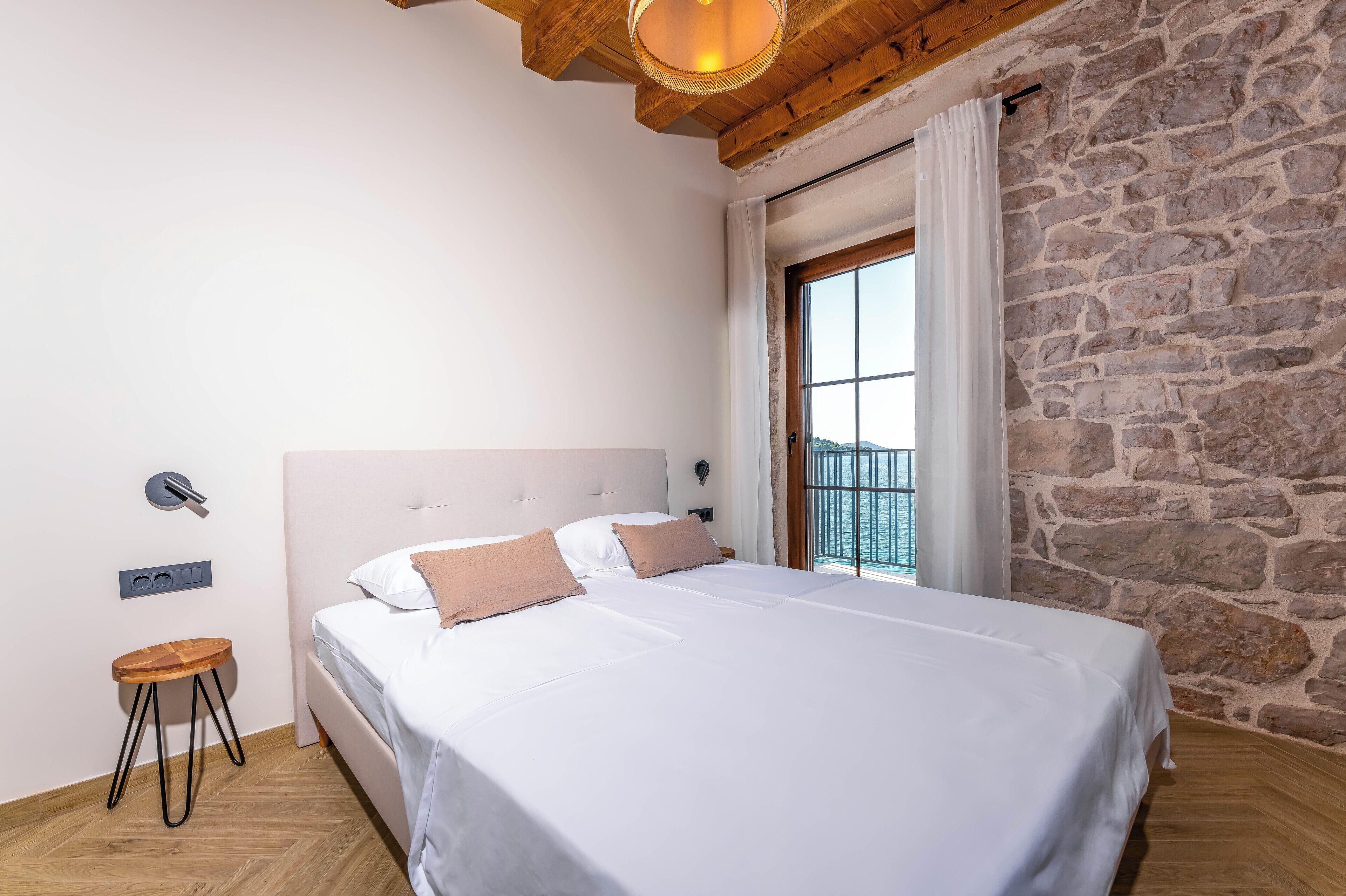A bedroom with a large bed, a wooden stool, a stone wall, and a window with a view.