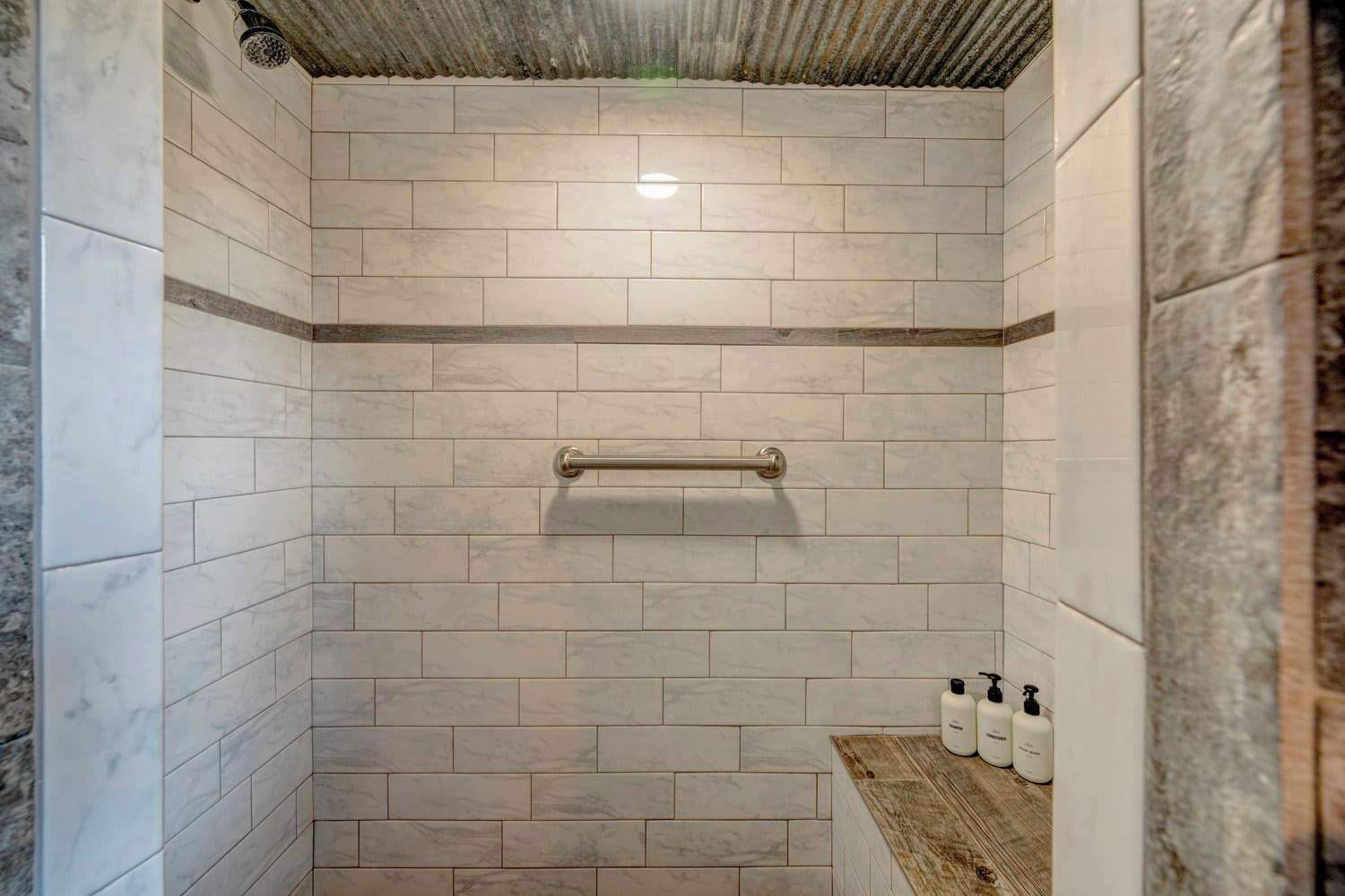 Una doccia con una barra di sostegno in metallo, piastrelle di marmo bianco e uno scaffale in legno con due bottiglie.