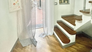Una habitación con escalera de madera, pared blanca con un estante pequeño y una puerta cubierta por cortinas que da a un espacio exterior.