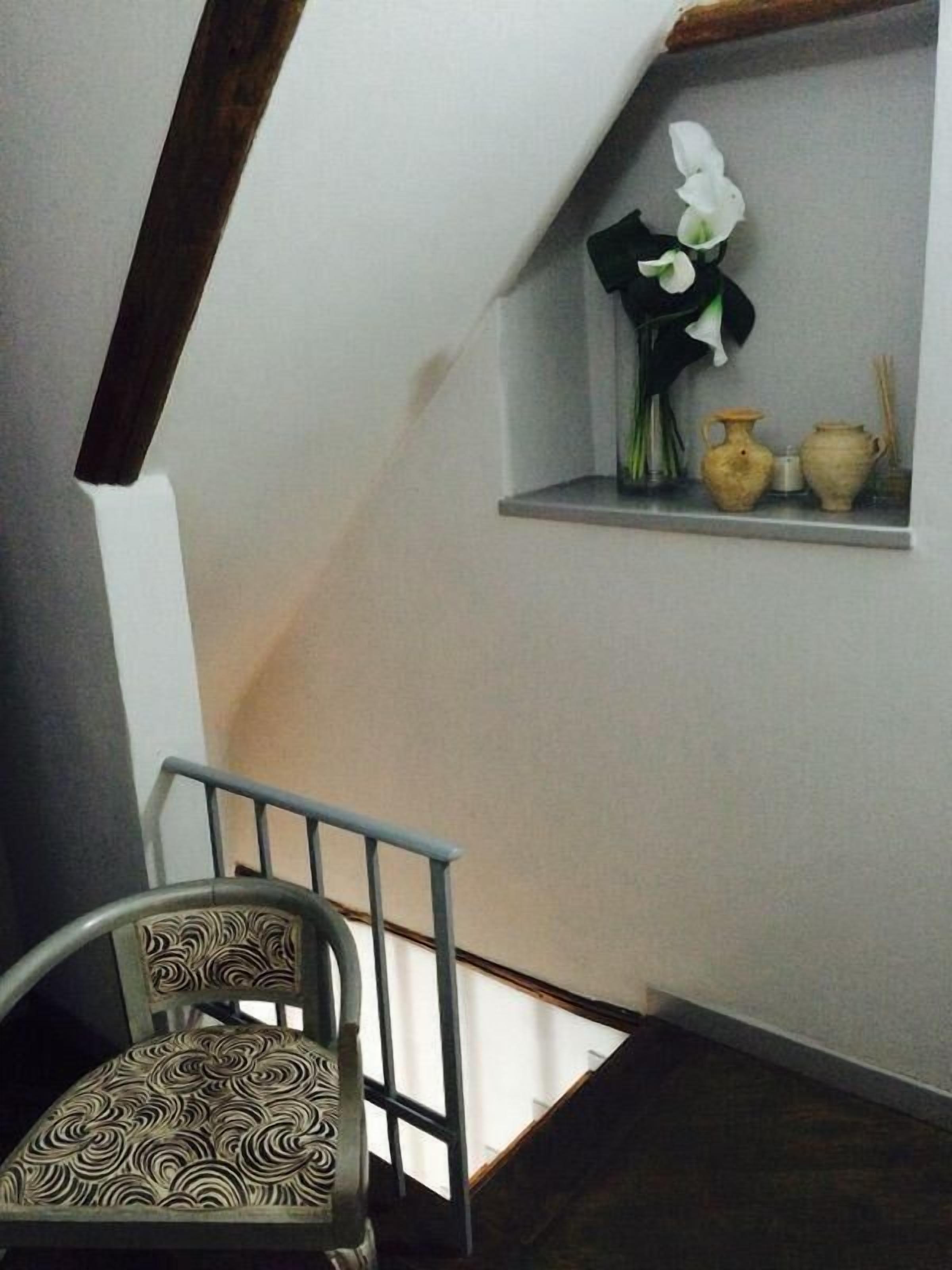 Una escalera con una silla al pie, un estante con adornos, y una pared blanca.