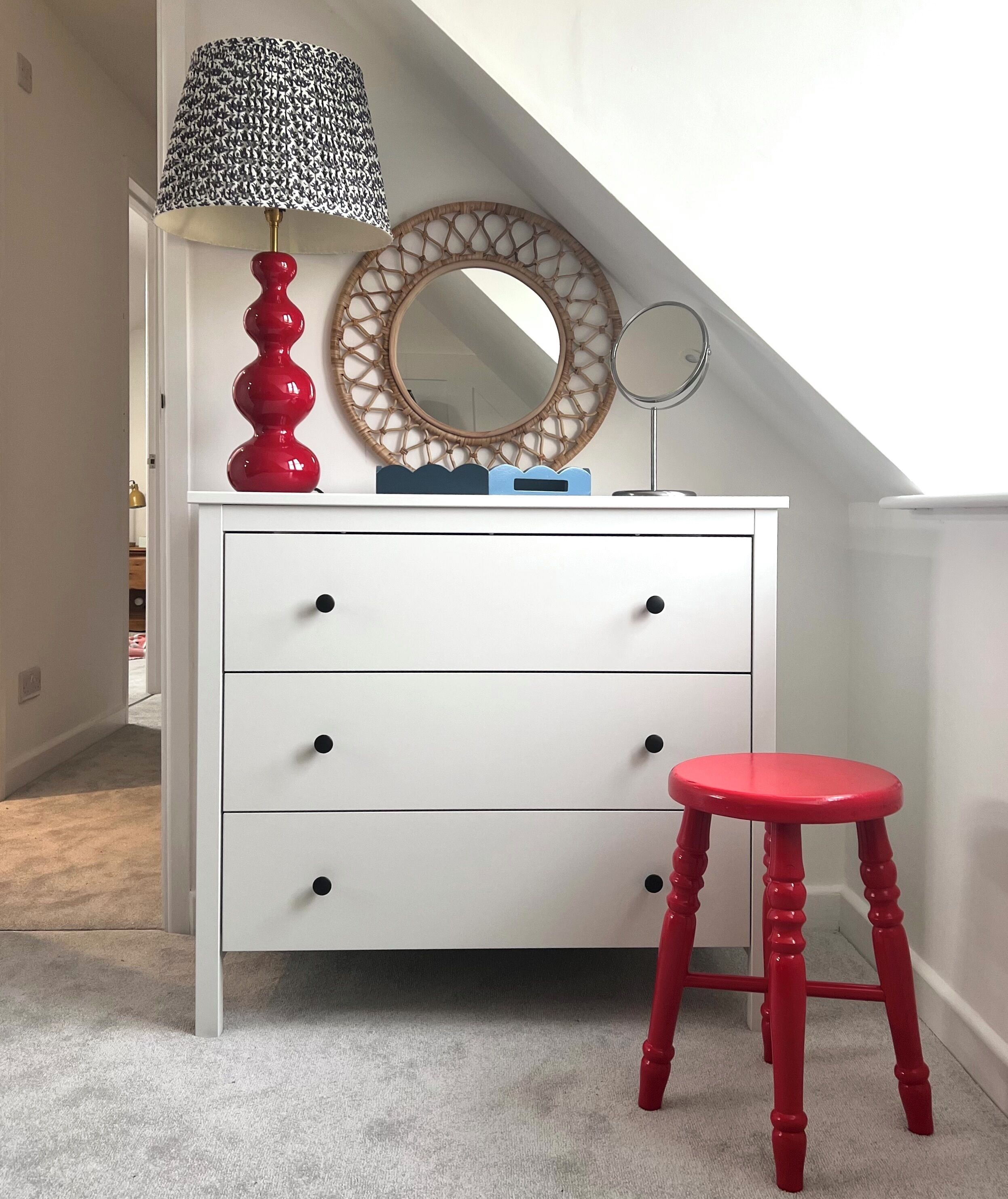 A white dresser with three drawers, a red stool, a lamp, and a round mirror with a wooden frame.
