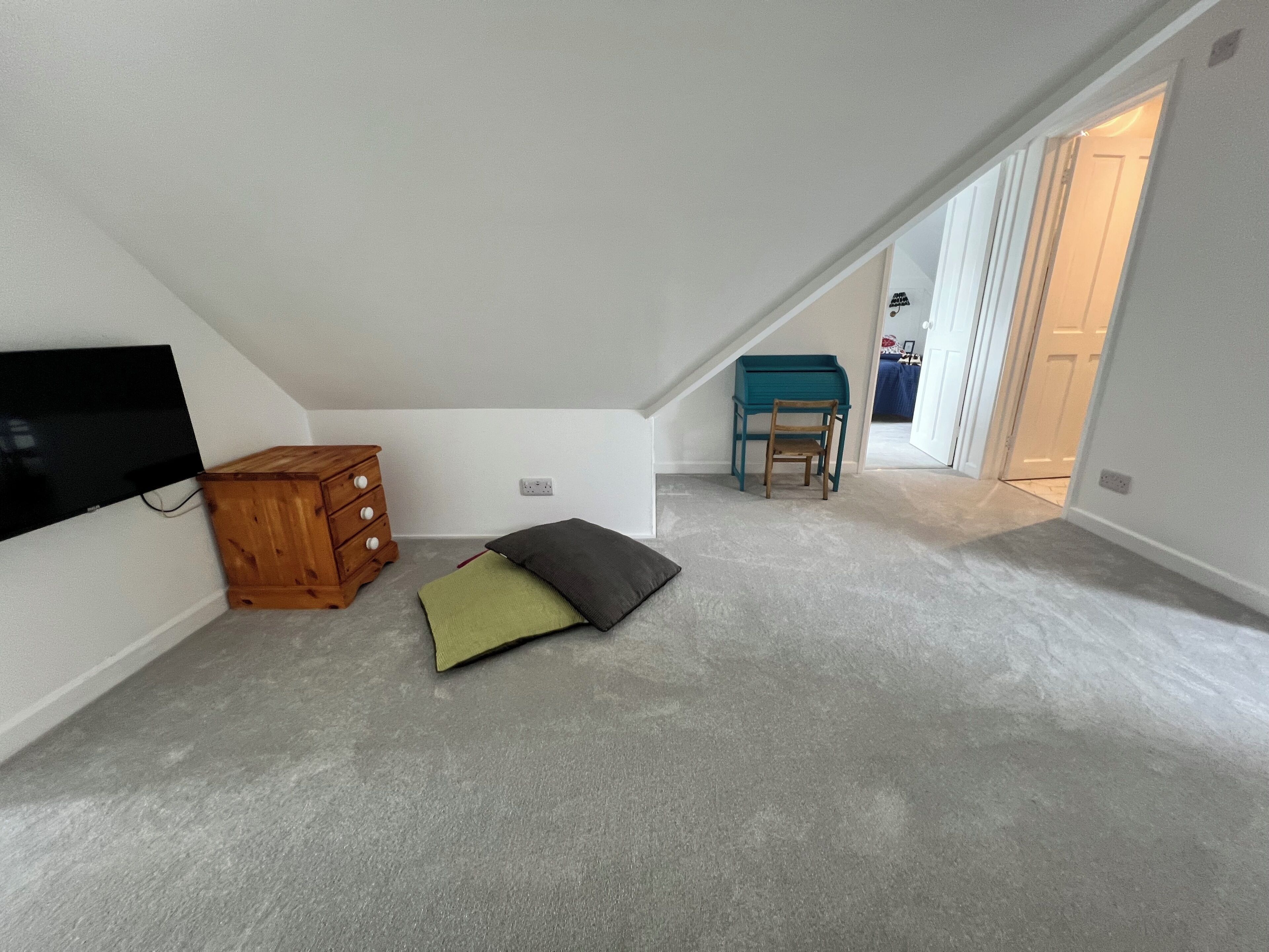 An attic room with a TV mounted on the wall, a wooden chest of drawers, a teal desk with a chair, and a carpeted floor.