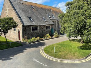 A stone building with a sloped roof, a paved road leading to it, and a grassy area with trees. - Beautiful cottage Snugglers Nook in heart of Bude near the beach, river & canal. (Bude)
