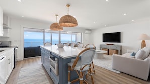 A modern kitchen with a large island, dining area with wicker chairs, a sofa, and a TV. - Coastal Inspired Three Bedroom Oceanfront Condo (Ocean City)