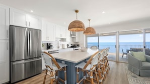 A modern kitchen with a large island, stainless steel appliances, wicker pendant lights, and a view of the ocean. - Coastal Inspired Three Bedroom Oceanfront Condo (Ocean City)
