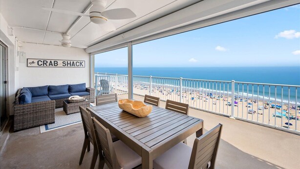 A balcony with a view of the beach, a table with chairs, a sofa, and a ceiling fan. - Coastal Inspired Three Bedroom Oceanfront Condo (Ocean City)