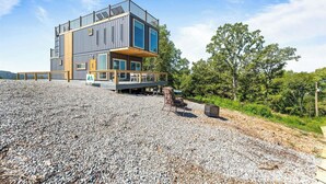 Ein modernes, mehrgeschossiges Haus mit Balkon, auf einer Schotterfläche mit Bäumen im Hintergrund gelegen.