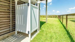 A white metal gate attached to a wooden structure with a pergola above, leading to a grassy area with a fence.