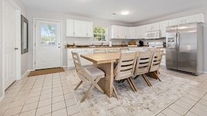 Une cuisine moderne avec des meubles blancs, un réfrigérateur en acier inoxydable, une table à manger en bois avec des chaises, et une grande fenêtre qui laisse entrer la lumière naturelle.