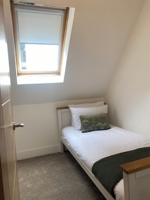 Quarto pequeno com uma cama de solteiro, teto inclinado, claraboia e porta com puxador metálico.
