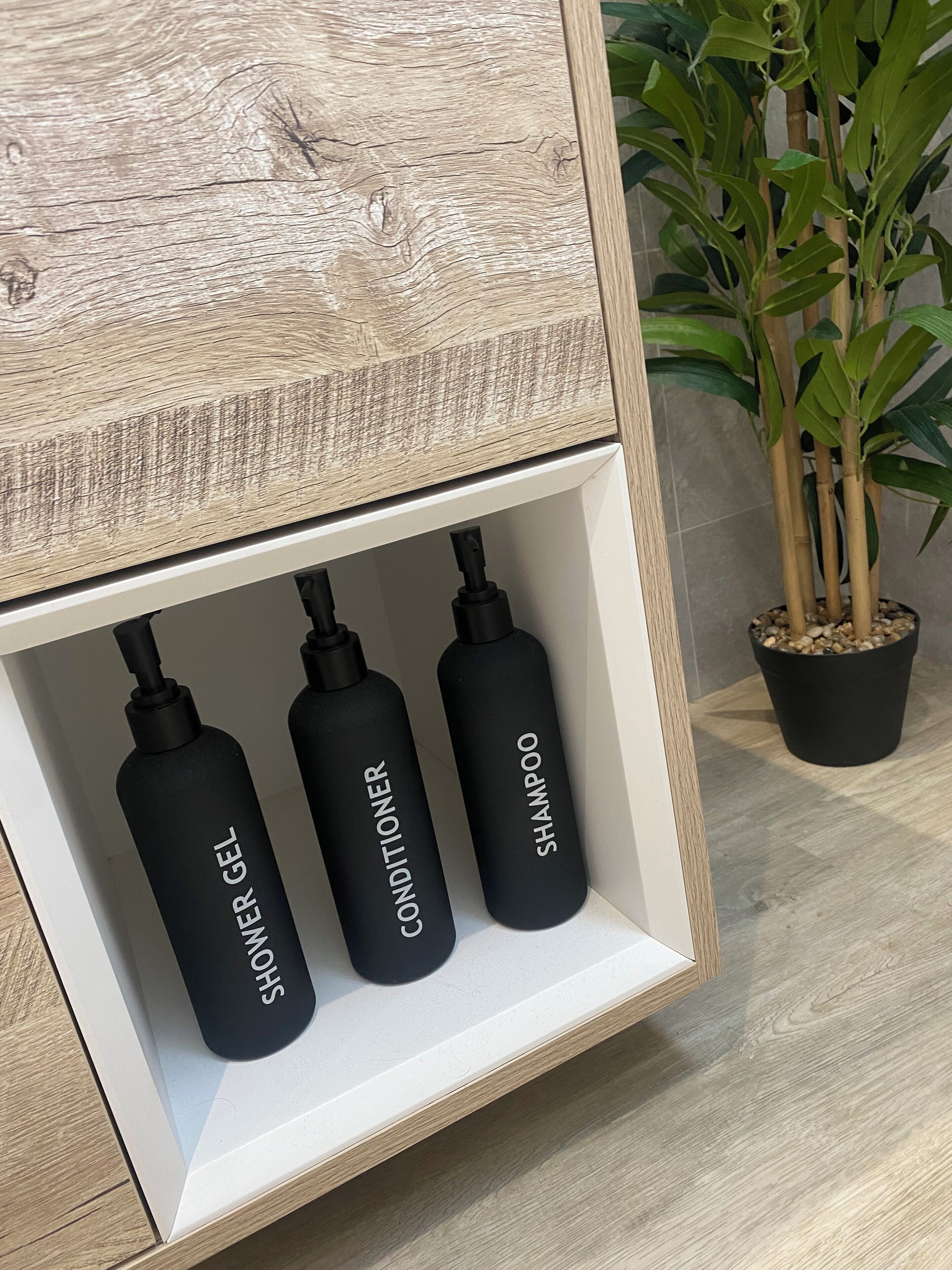 A wooden shelf with three black bottles labeled "Shower Gel," "Conditioner," and "Shampoo."