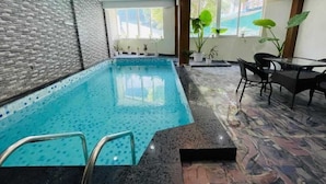 Una piscina interior con suelo de baldosas, pared de ladrillo y zona de estar con sillas y una mesa.