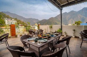 A terrace with wicker furniture and a table set for a meal, overlooking a mountainous landscape. - The Shore By Blue Waves (Narendranagar)