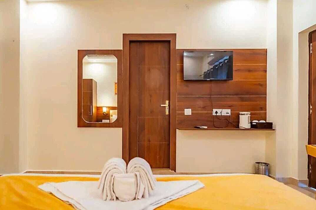 A hotel room with a bed, a wooden door, a mounted TV, and a small shelf with toiletries.